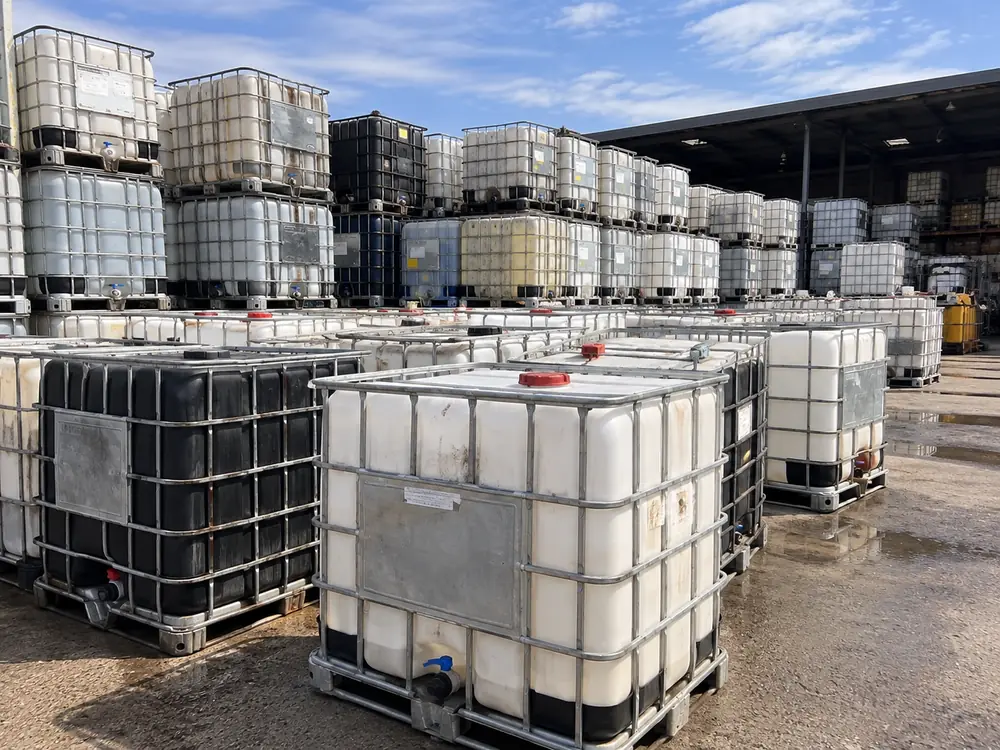 A mixed lot of used IBC totes of varying condition stacked in a yard awaiting collection